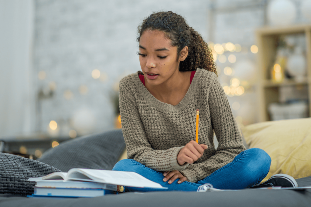 A student sits on a bed, reading a book while holding a pencil
